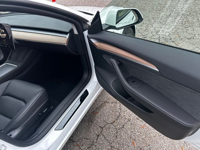 Open door of a white Tesla Model 3. Interior features black seats, door panel, and silver trim.