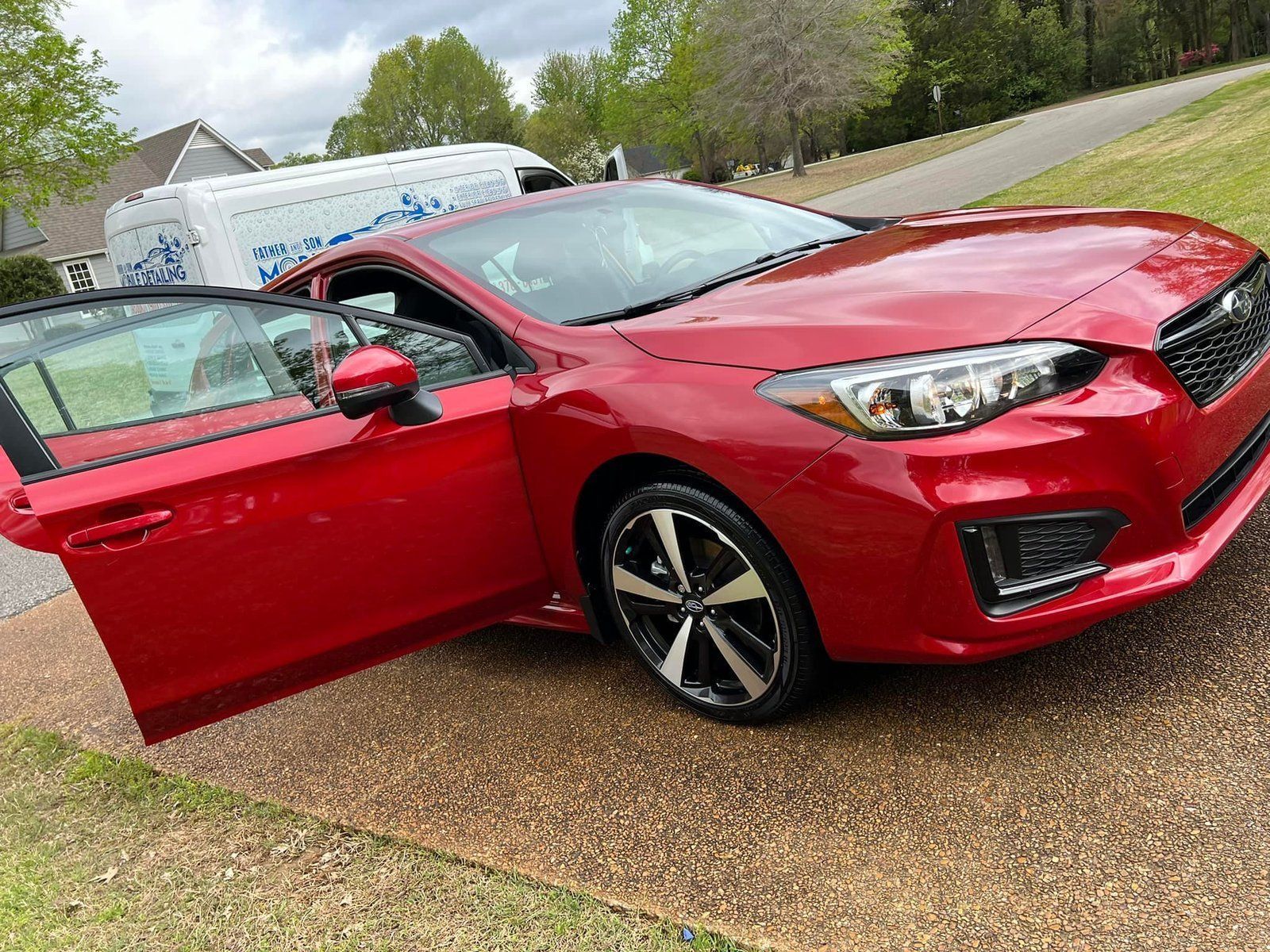 Red Subaru Impreza with open door parked on a driveway, service van in the background.