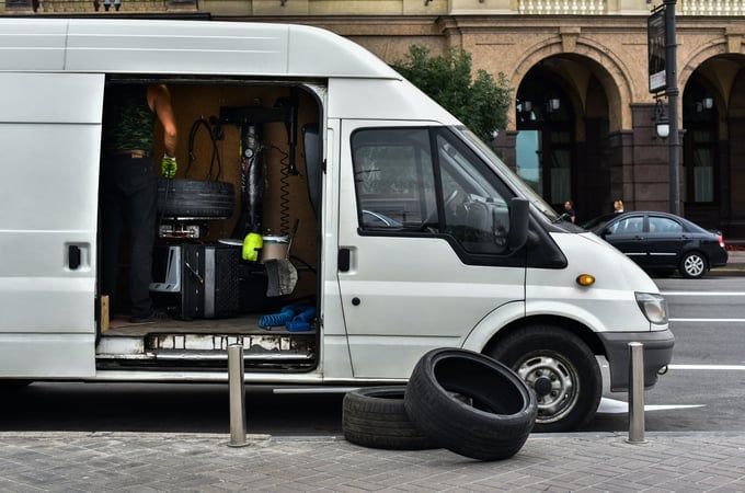 White van with open side door, tires on the ground, person inside working on something on a city street.