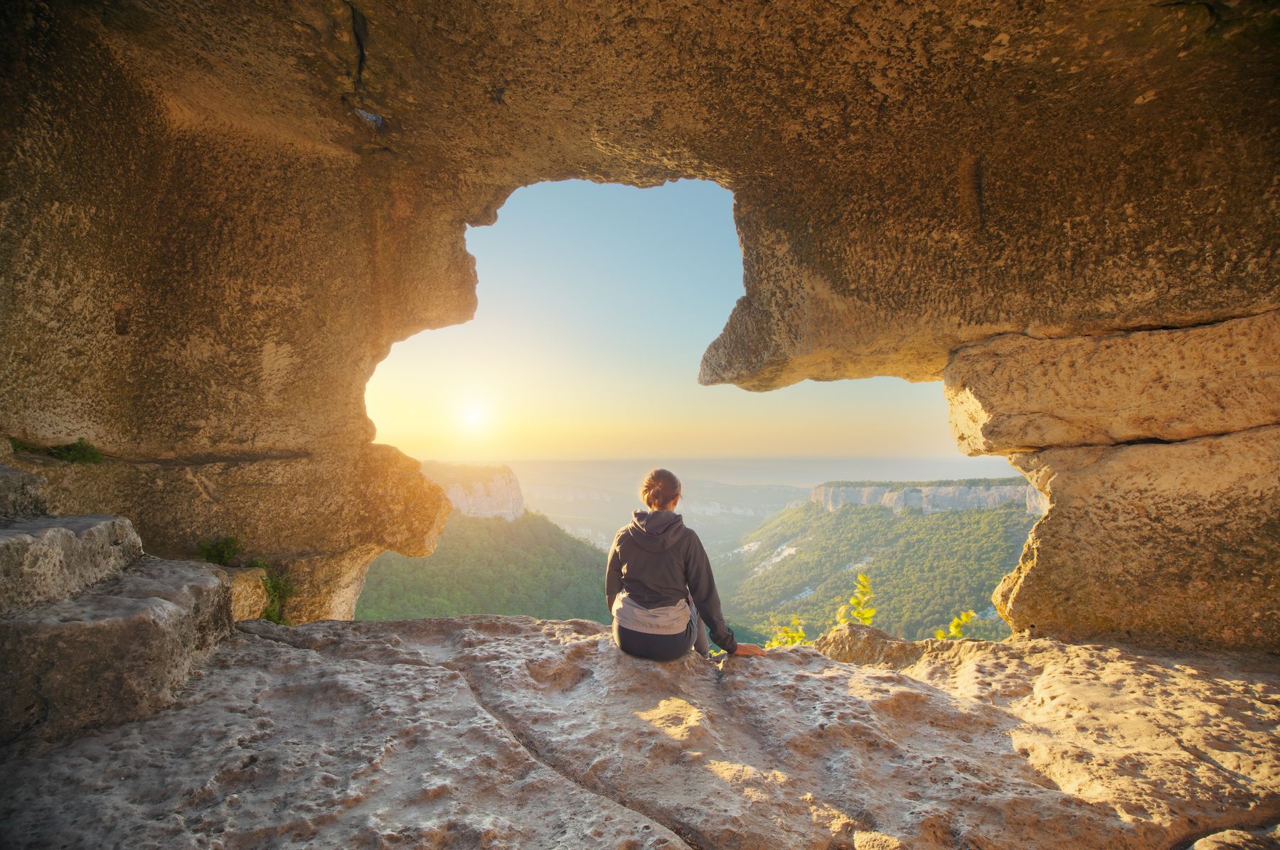 A man is sitting in a cave looking at the sunset.