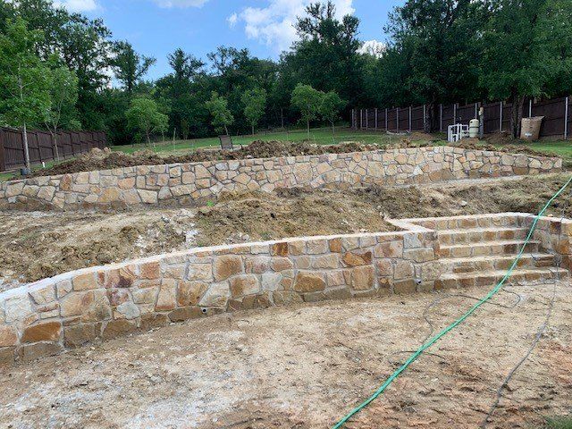 Stone retaining walls creating tiered levels in a backyard, with a brown fence and trees in the background.