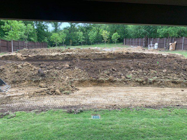 Backyard with dirt mounds, likely construction. Green grass, brown fence, trees.