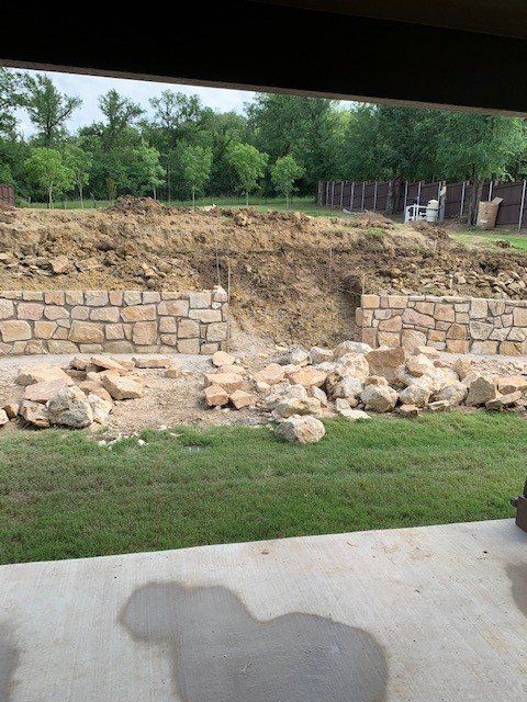 Stone retaining wall construction in a yard, with dirt mound and scattered rocks, trees in the background.