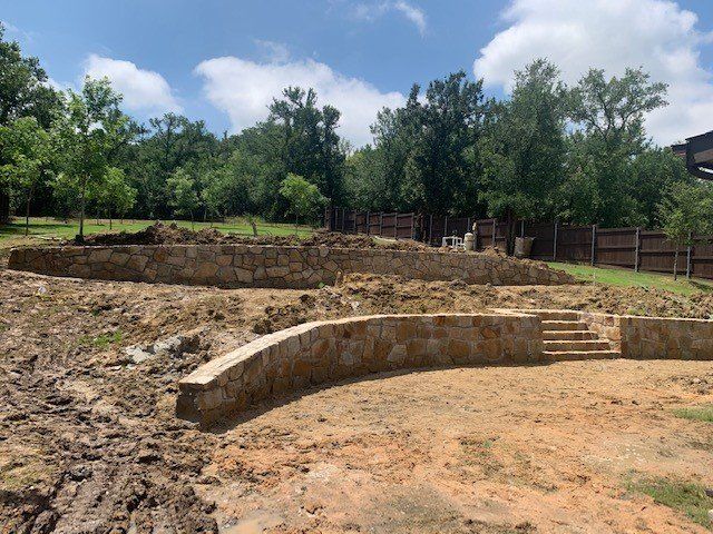 Stone retaining walls under construction in a backyard with steps, dirt, and trees.