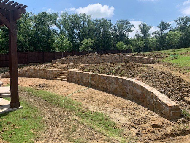 Stone retaining walls on a sloped yard, with steps and dirt, trees in the background.