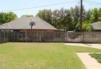 A backyard with a wooden fence and a basketball hoop, surrounded by grass and trees.