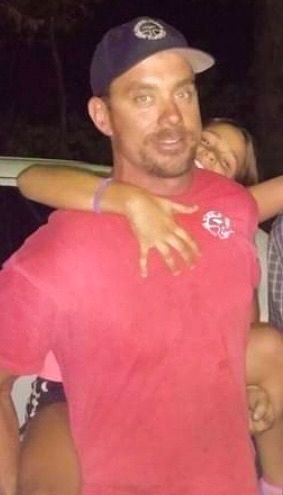 A man in a red shirt and hat is holding a little girl on his shoulders.