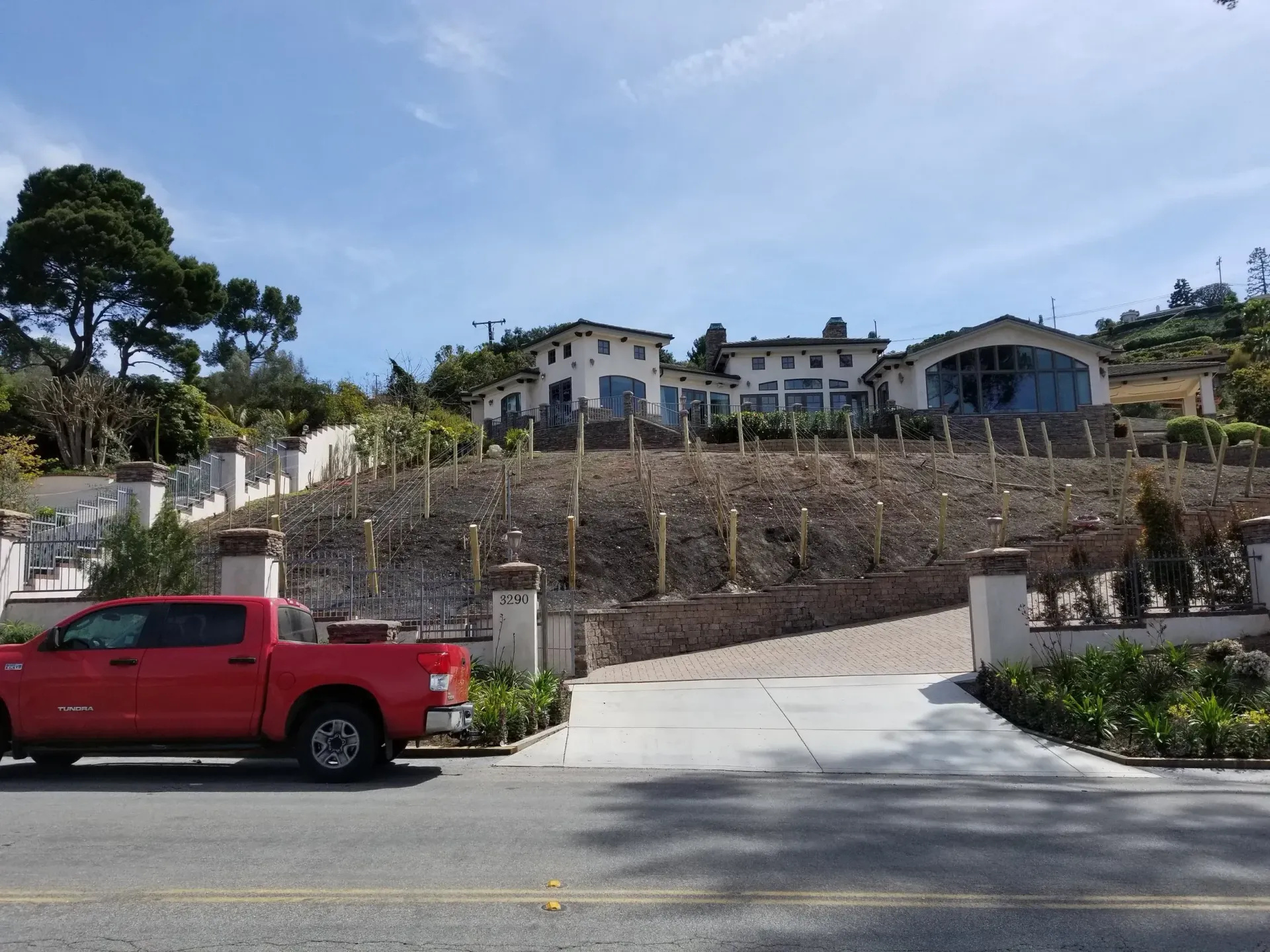 Red truck parked on a road in front of a large house on a hill with vineyard trellises; blue sky.