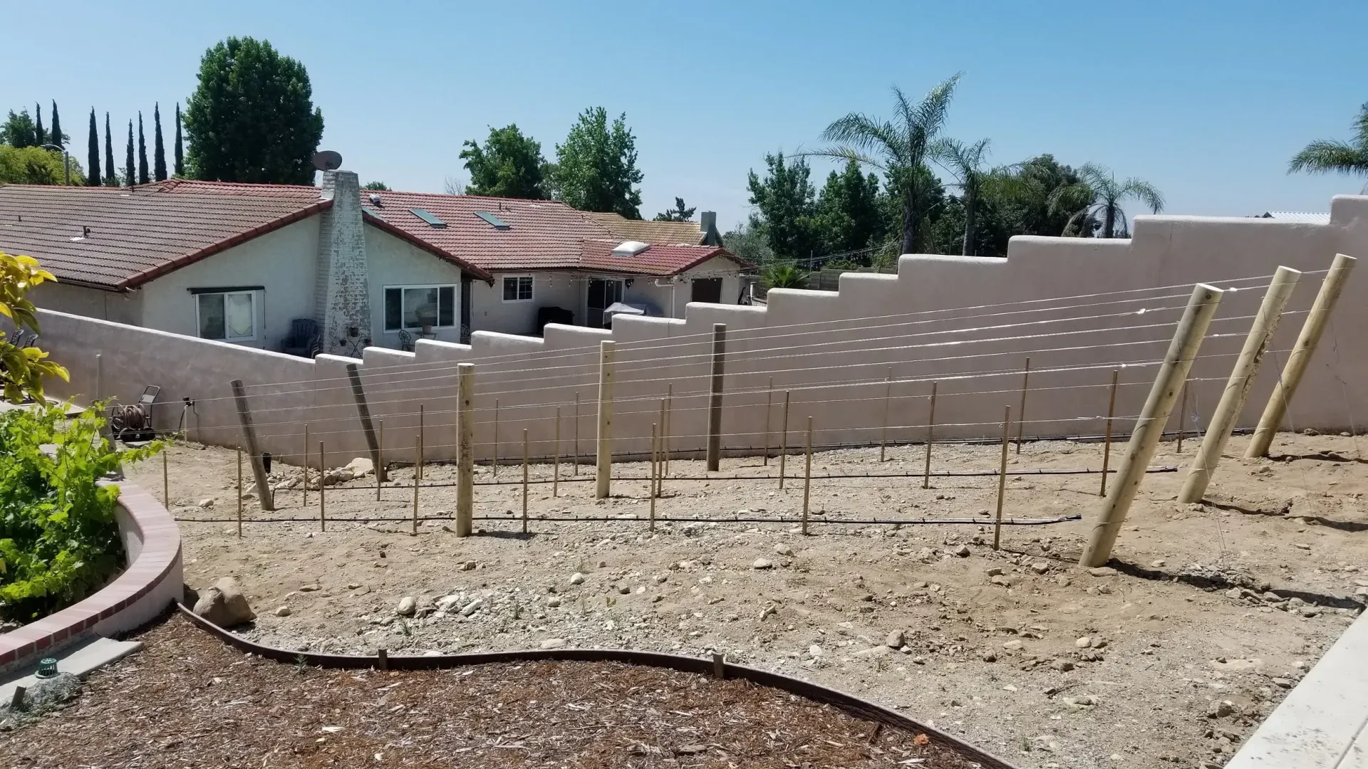 Backyard with dirt and rocks, wooden posts, a textured wall, and a house in the background on a sunny day.