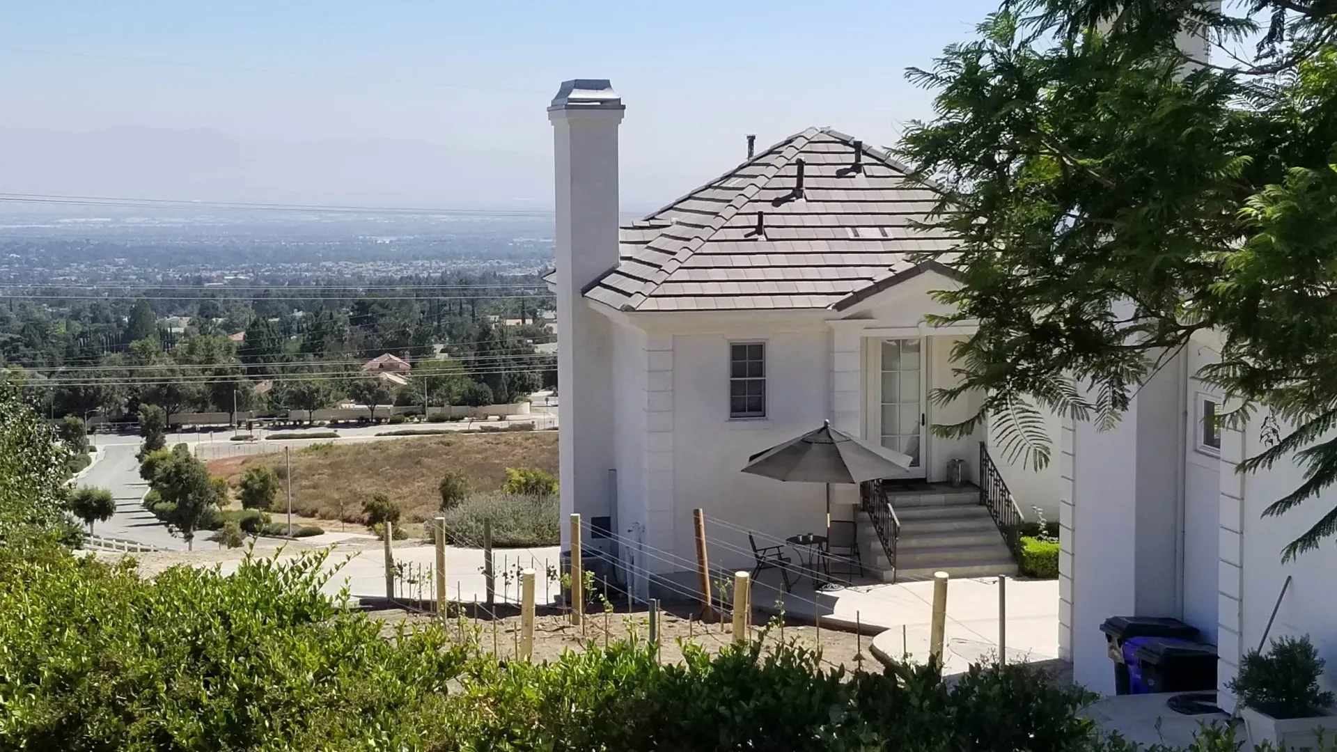 White house with chimney, patio, and steps, overlooking a cityscape under a hazy, sunny sky.
