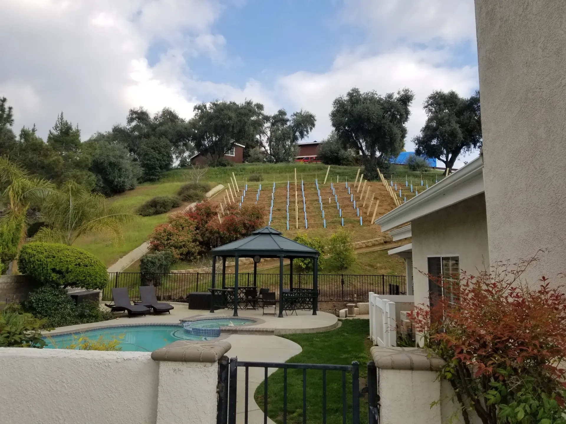 A backyard with a pool, gazebo, and hillside vineyard under a cloudy sky.