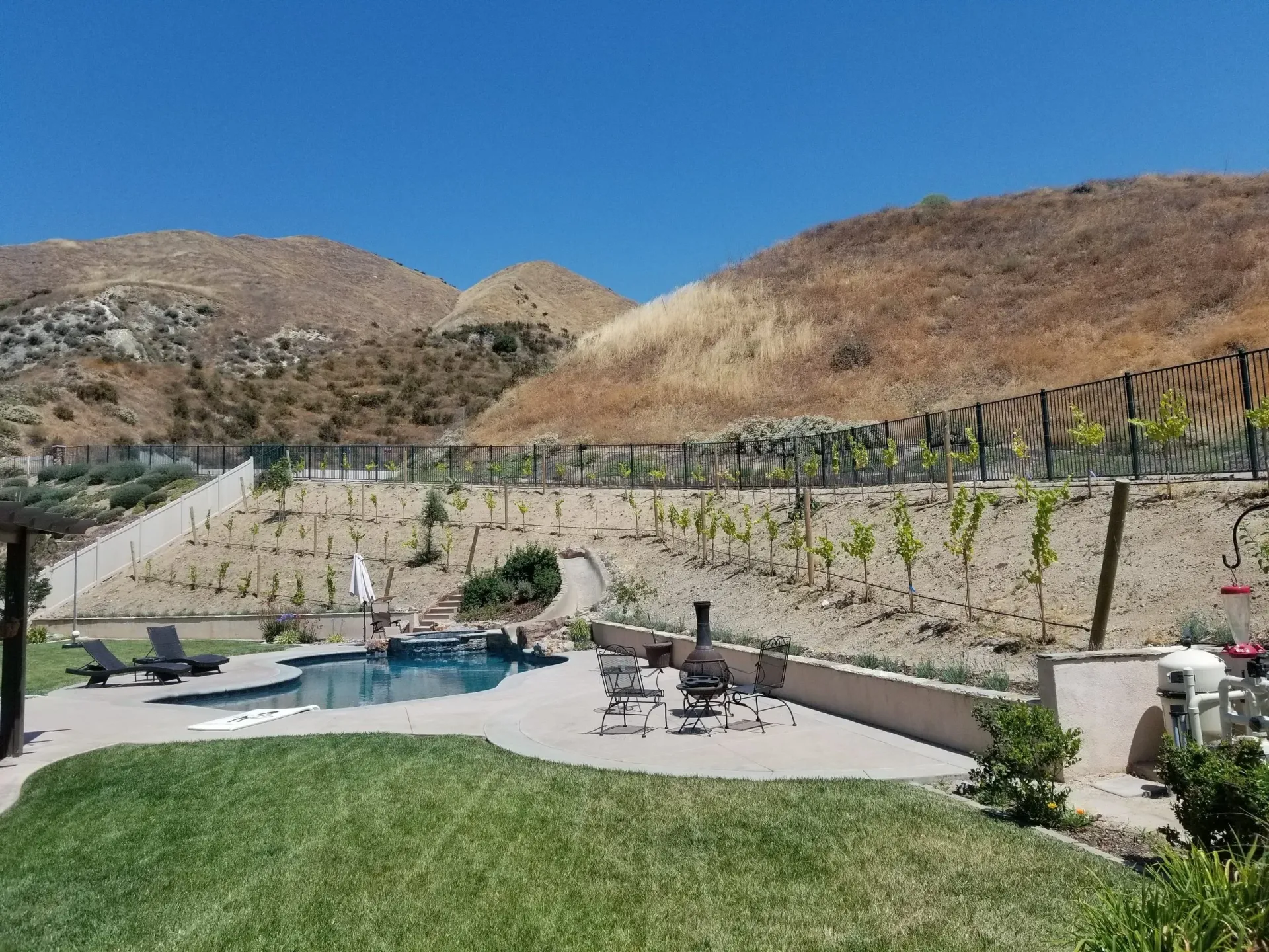 Swimming pool and patio overlooking dry hills, lush green grass in foreground. Blue sky.