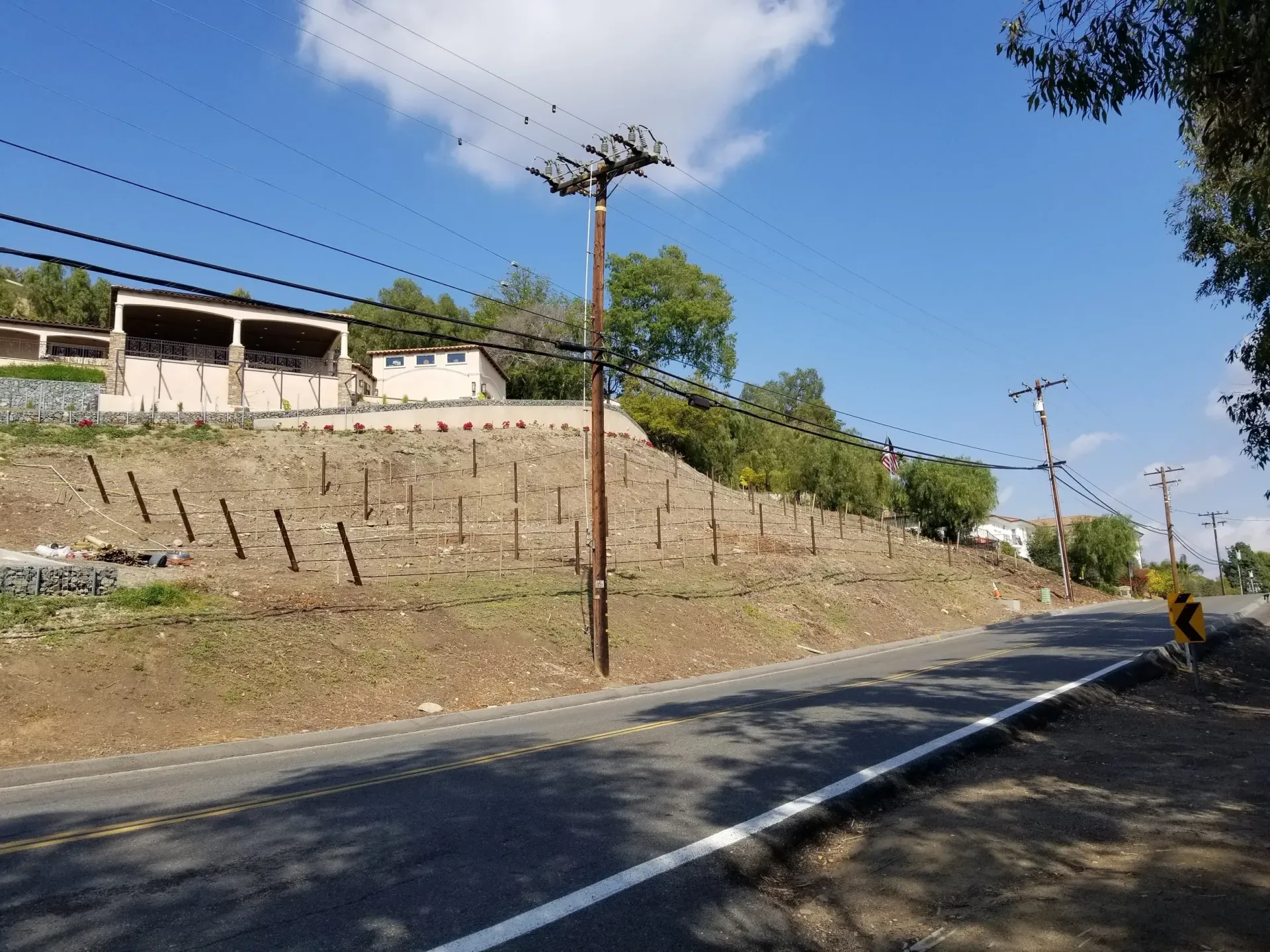 Roadside view with utility poles, hillside, and buildings under a blue sky with clouds.