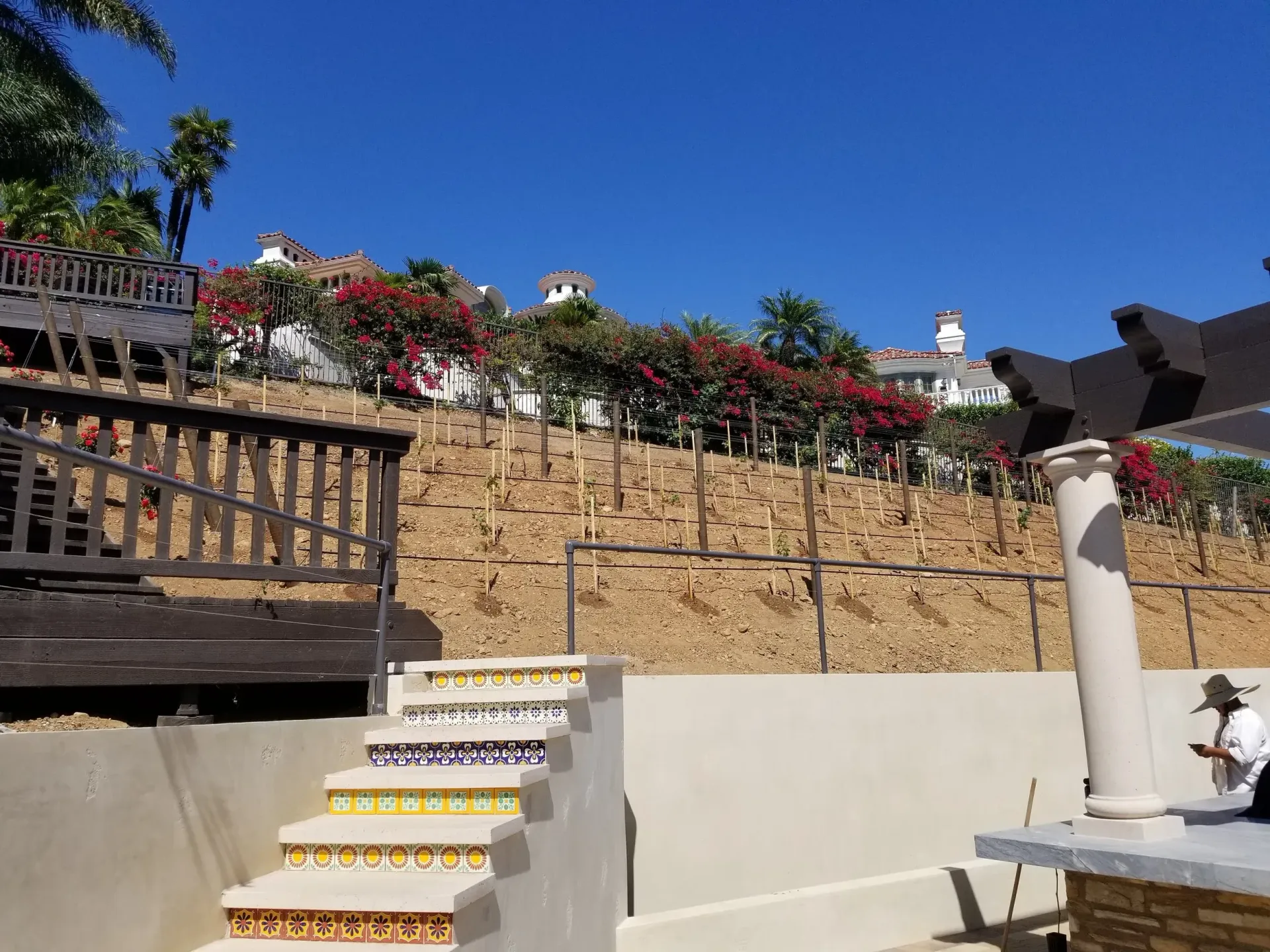 Staircase with colorful tiles leading up to a wooden deck and a hillside with red flowers and buildings under a blue sky.