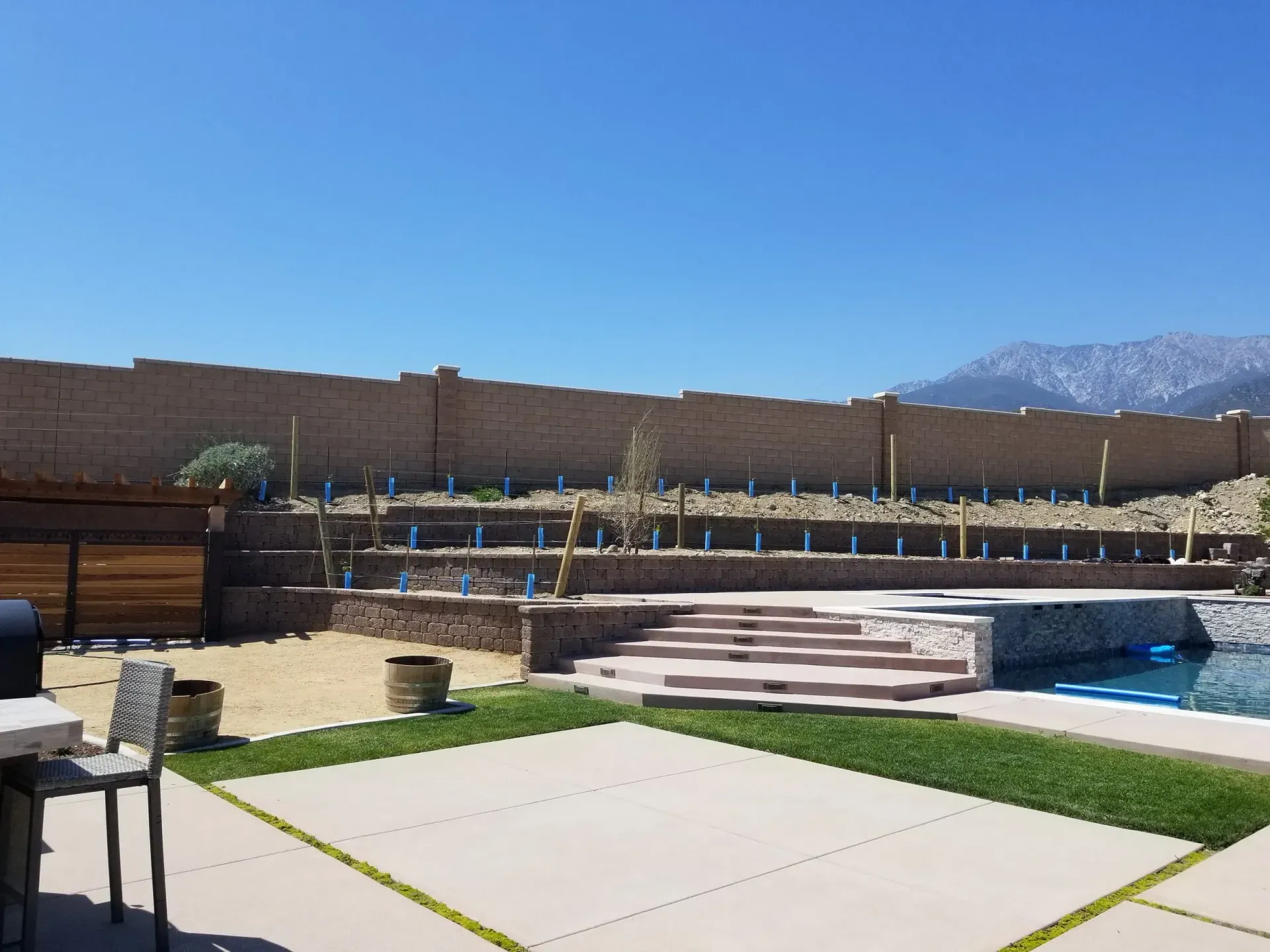 Backyard with pool, tiered retaining wall with plants, patio, and mountain view under blue sky.