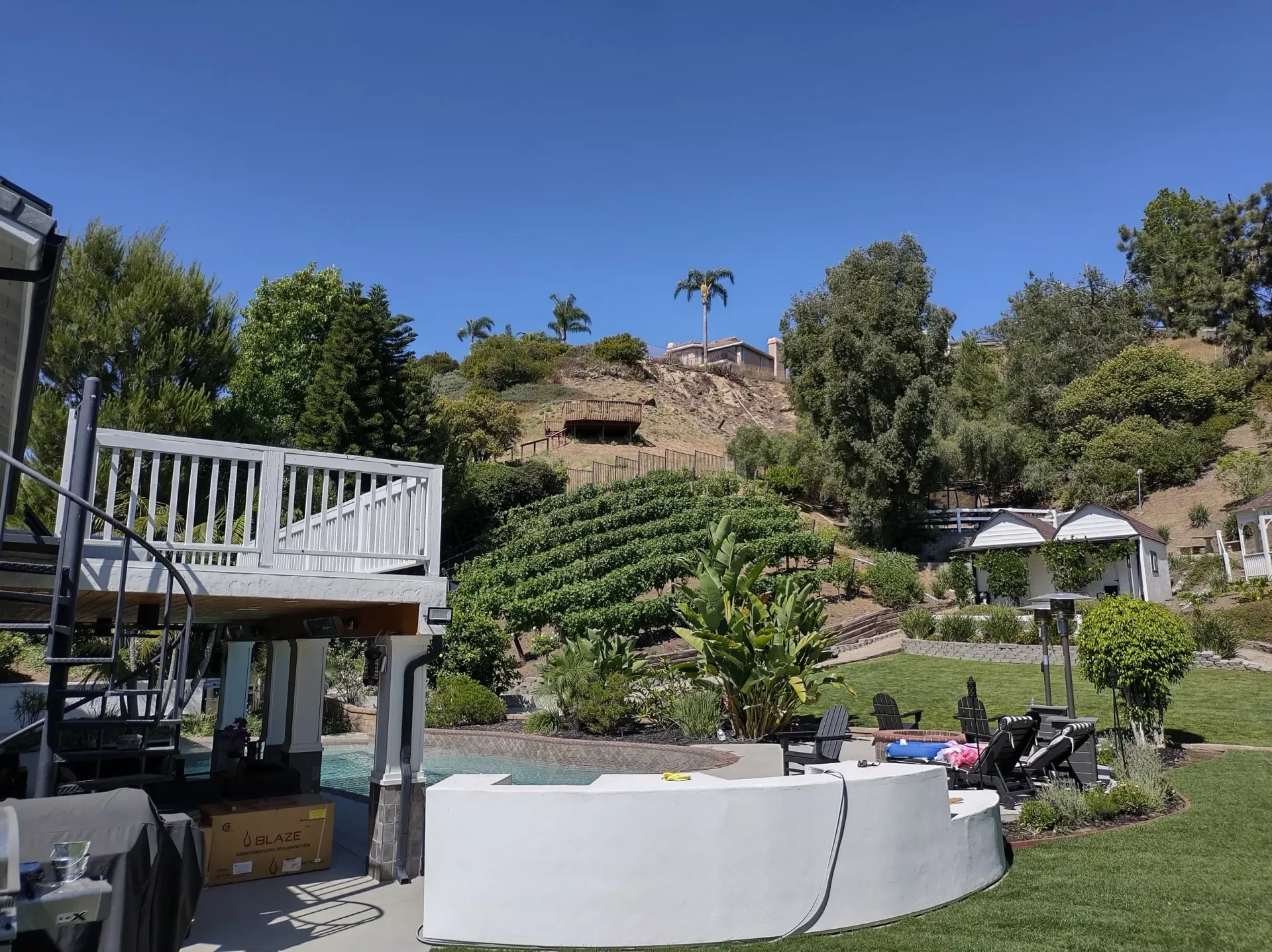 Backyard with pool, deck, and hillside with trees under a blue sky.