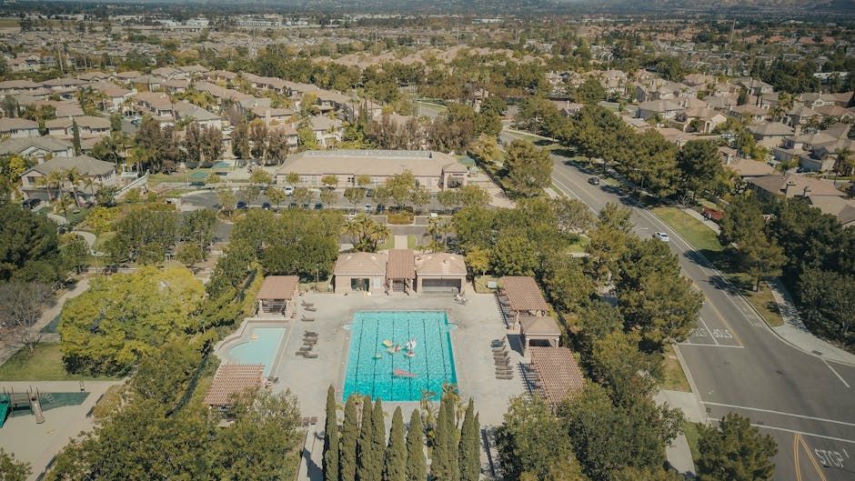 Aerial view of a residential swimming pool area surrounded by trees, sidewalks, and suburban homes.
