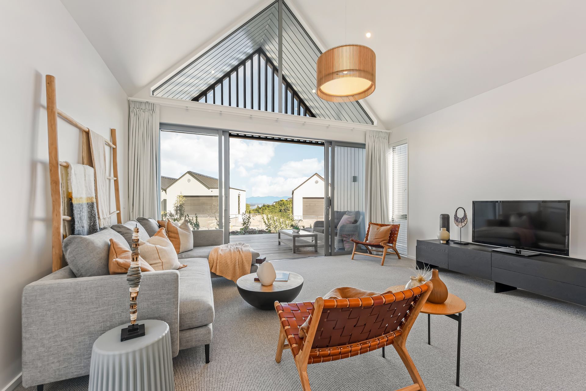Bright living room with open doors to a view. Gray sofa, wooden chair, coffee table, TV, copper light fixture.