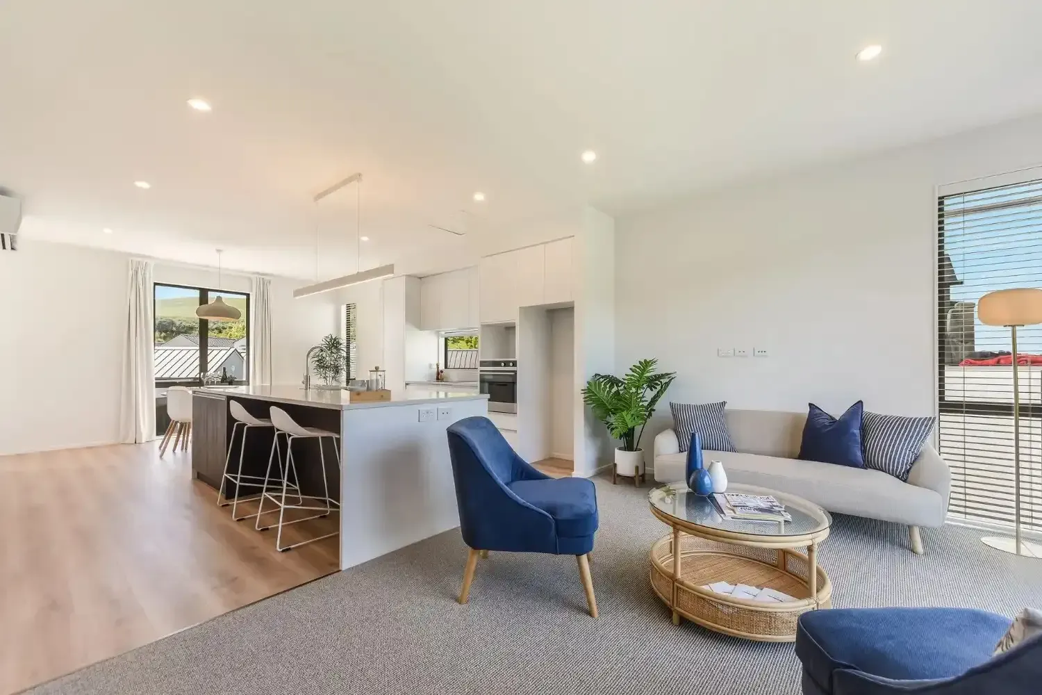 Modern living room with kitchen, light wood floors, blue accents, and large windows.