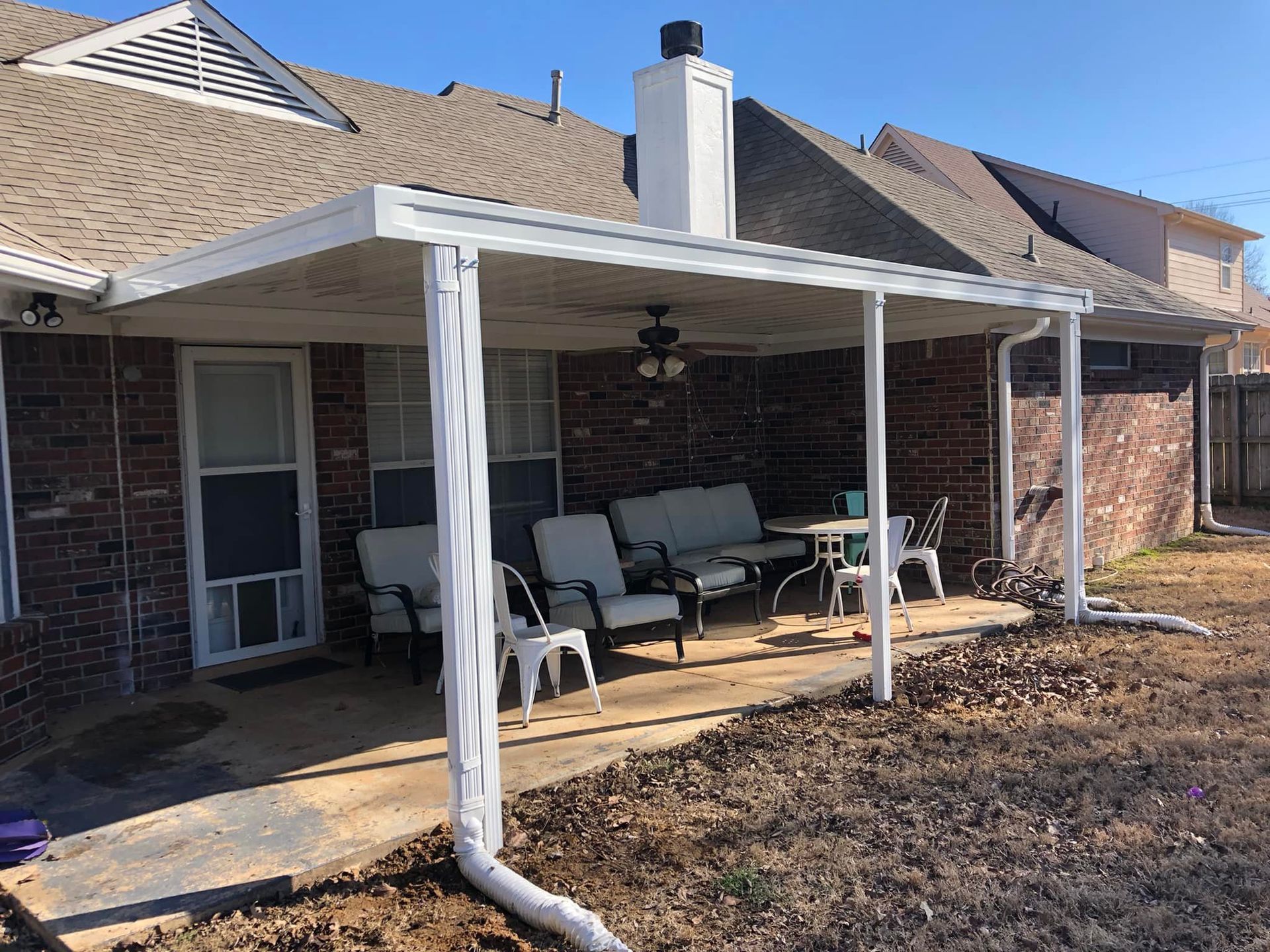A covered patio with a couch , chairs , table and a ceiling fan in front of a brick house.