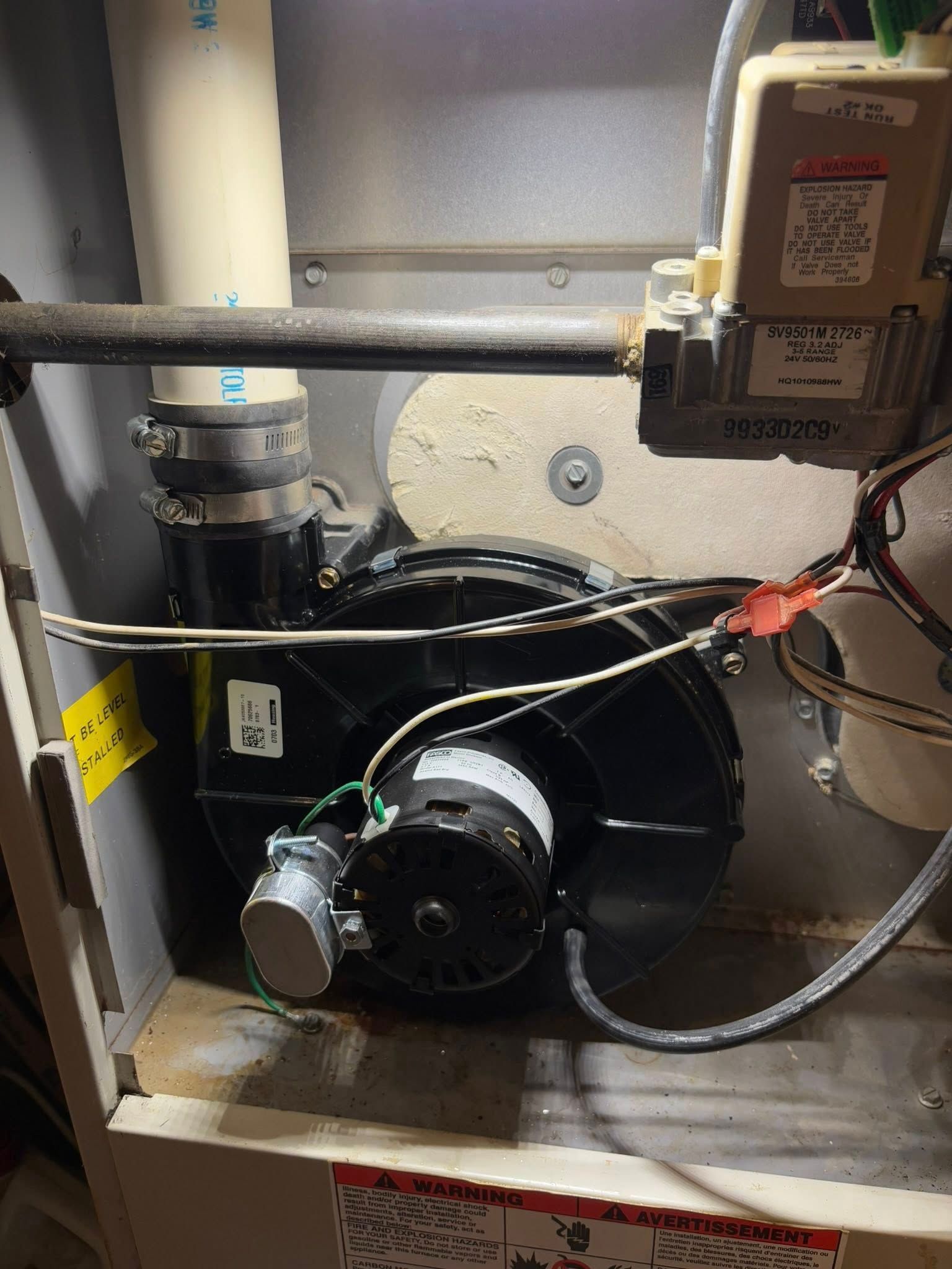 Furnace blower unit with black fan and motor, gas valve, and vent pipes.