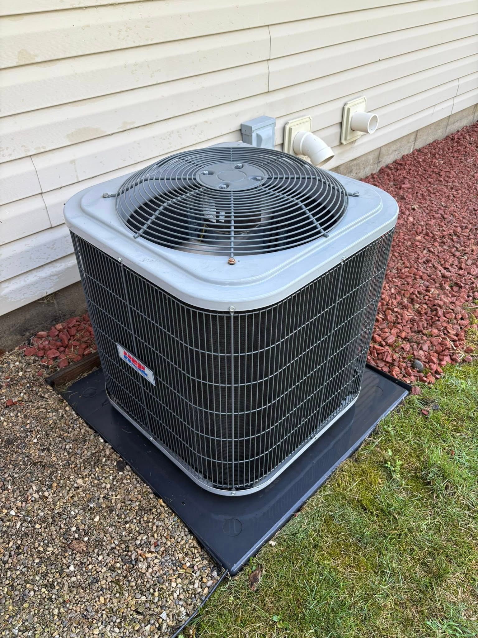 An air conditioning unit sits outside next to a building with red rocks and grass.