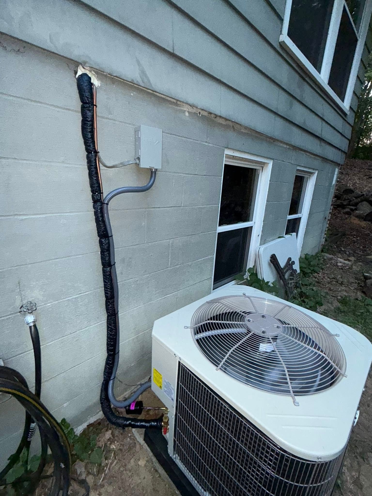 HVAC unit outside a concrete building. Black insulated tubes run up the wall to a gray box.