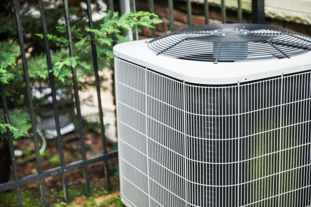 Air conditioning unit with fan, outdoors near a fence and foliage.