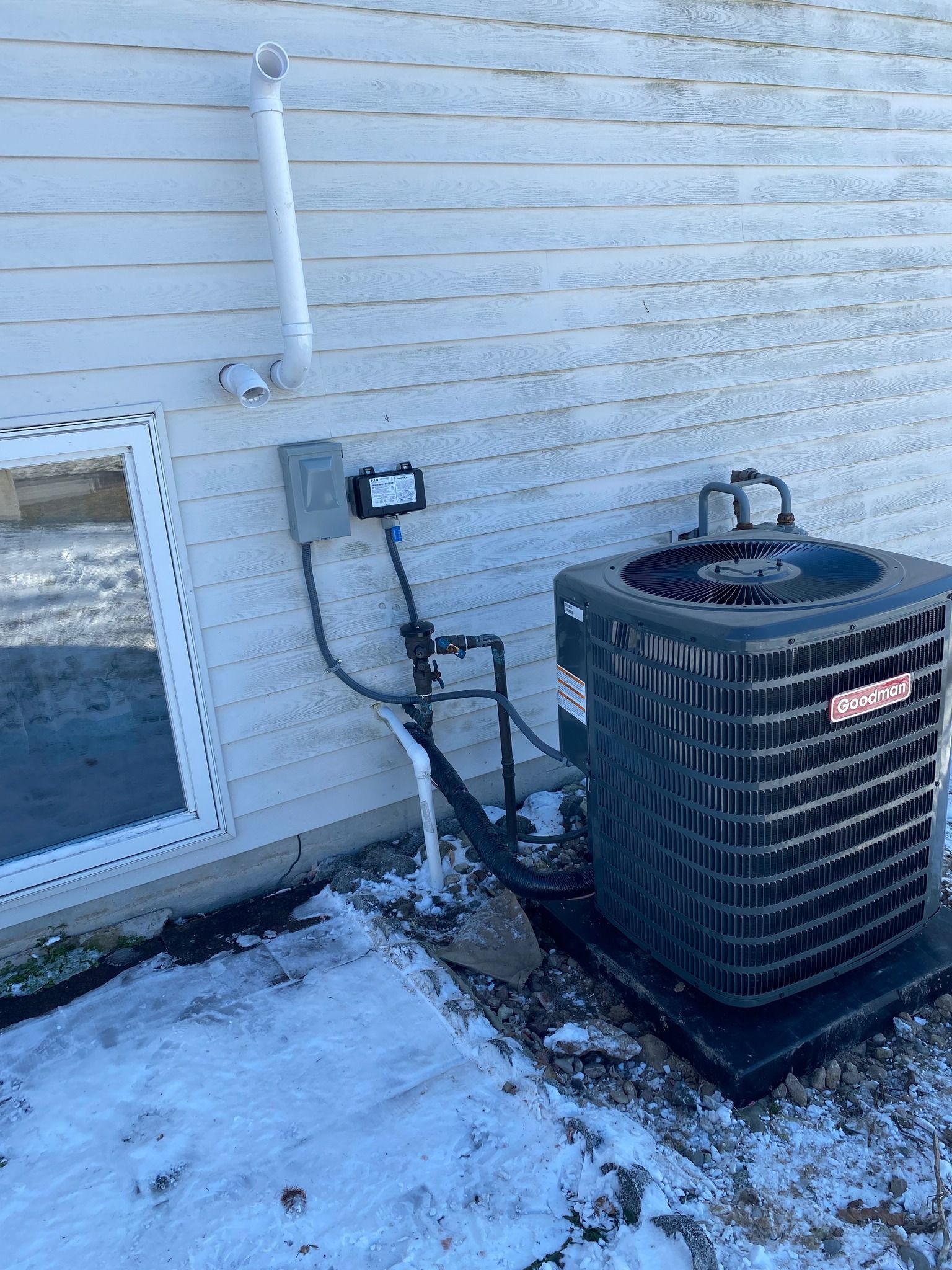 A dark grey Goodman air conditioning unit sits on a concrete pad against a white-sided house wall with electrical conduit.