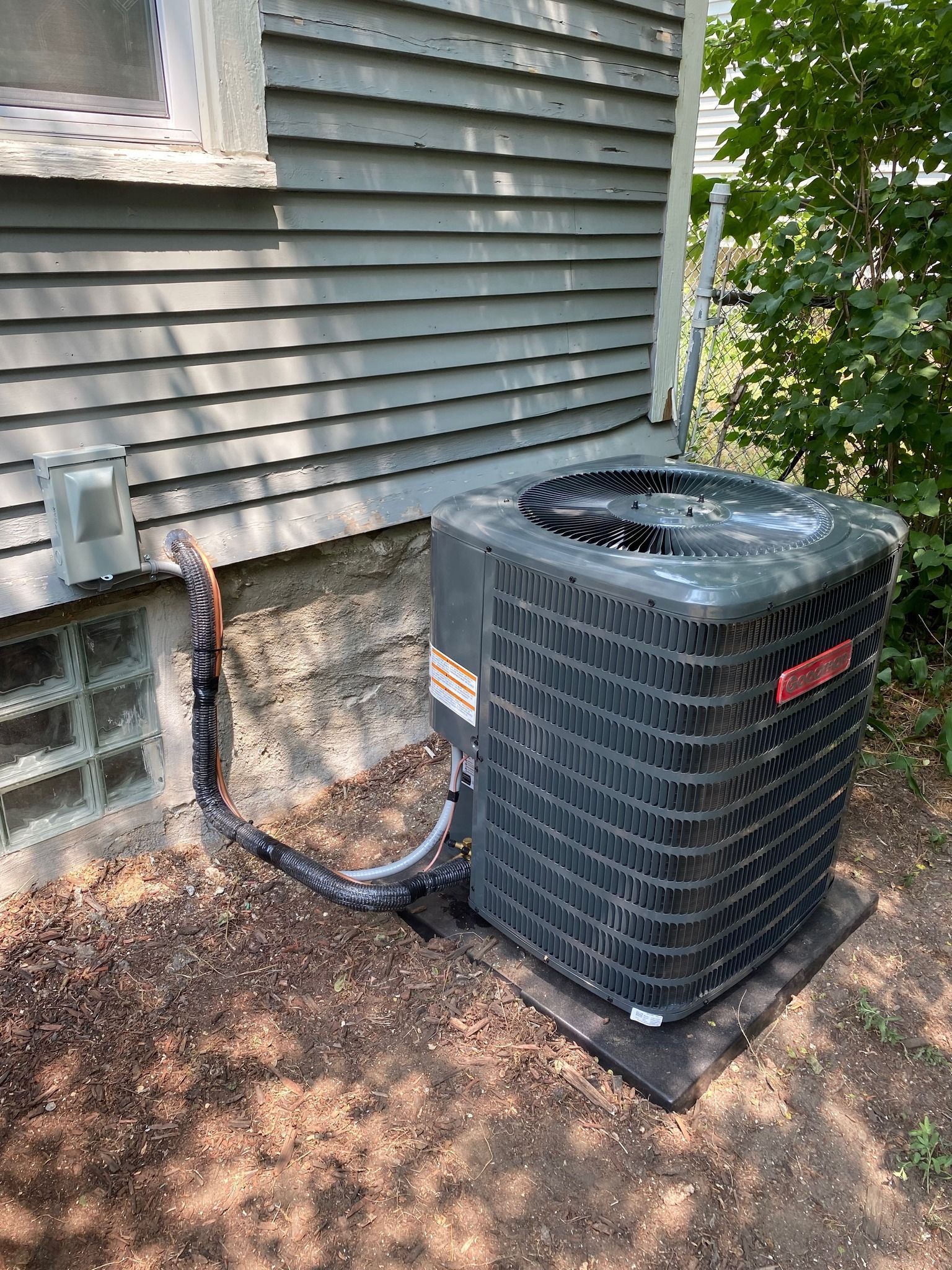 A dark grey Goodman air conditioning unit sits on a black pad next to a house with grey siding and a glass block window.