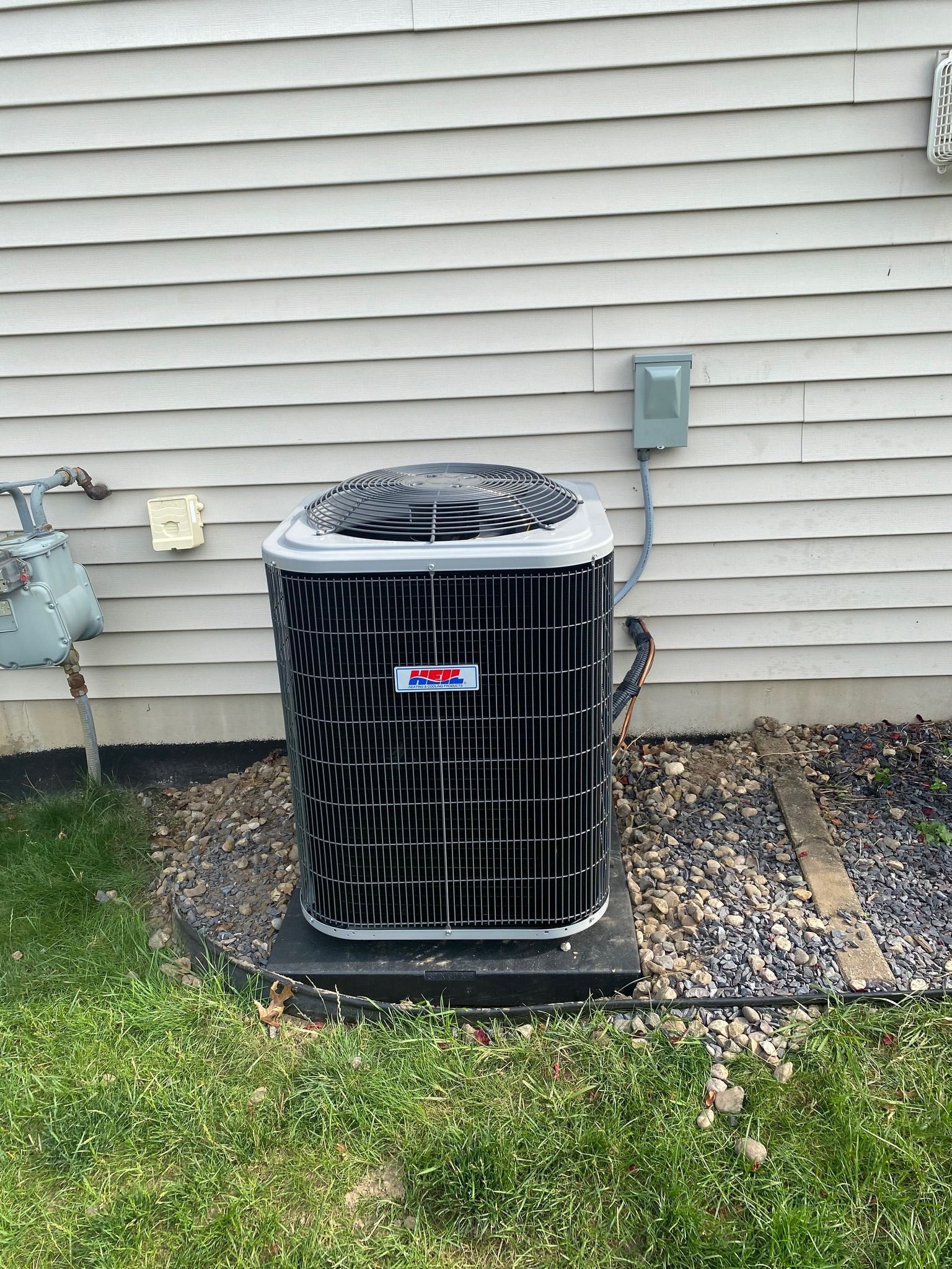 A grey HVAC outdoor condenser unit sitting on a concrete pad against white horizontal vinyl siding.