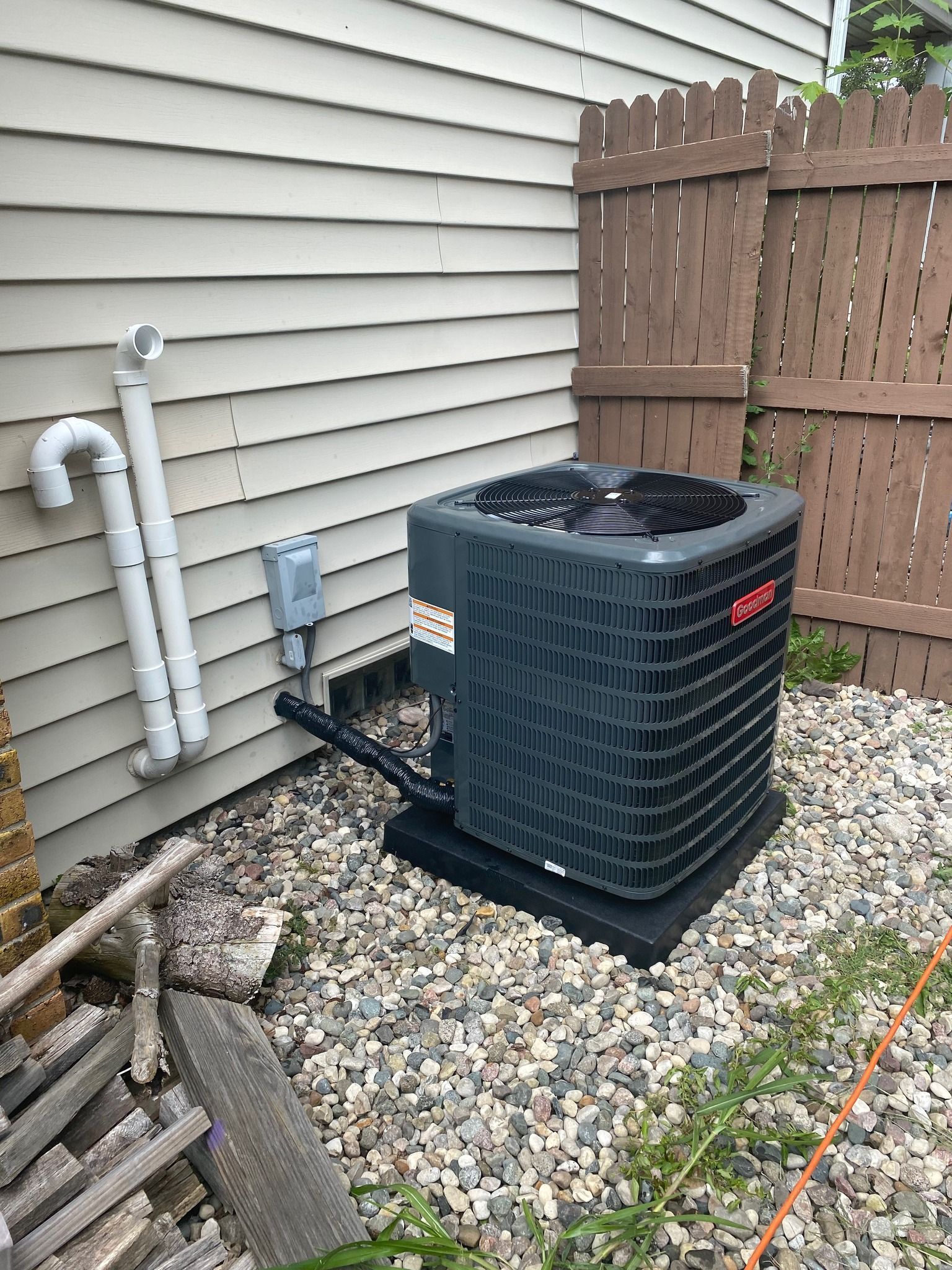 A grey air conditioning unit sits on a gravel patch beside a house exterior with white siding and two PVC pipes.