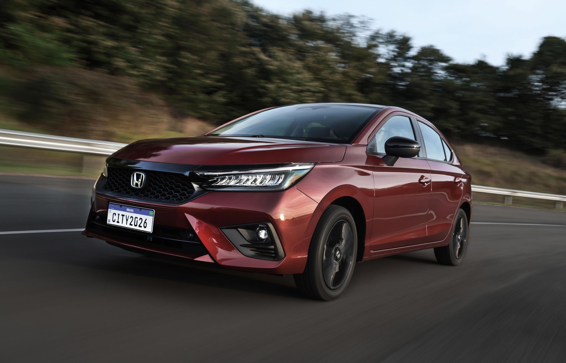 Sedan Honda City vermelho em uma rodovia, trafegando para a esquerda com árvores ao fundo.