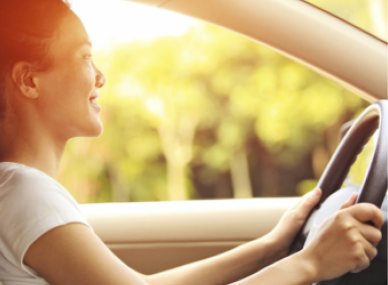 Mulher sorrindo enquanto dirige um carro, com as mãos no volante e a luz do sol brilhando pela janela.