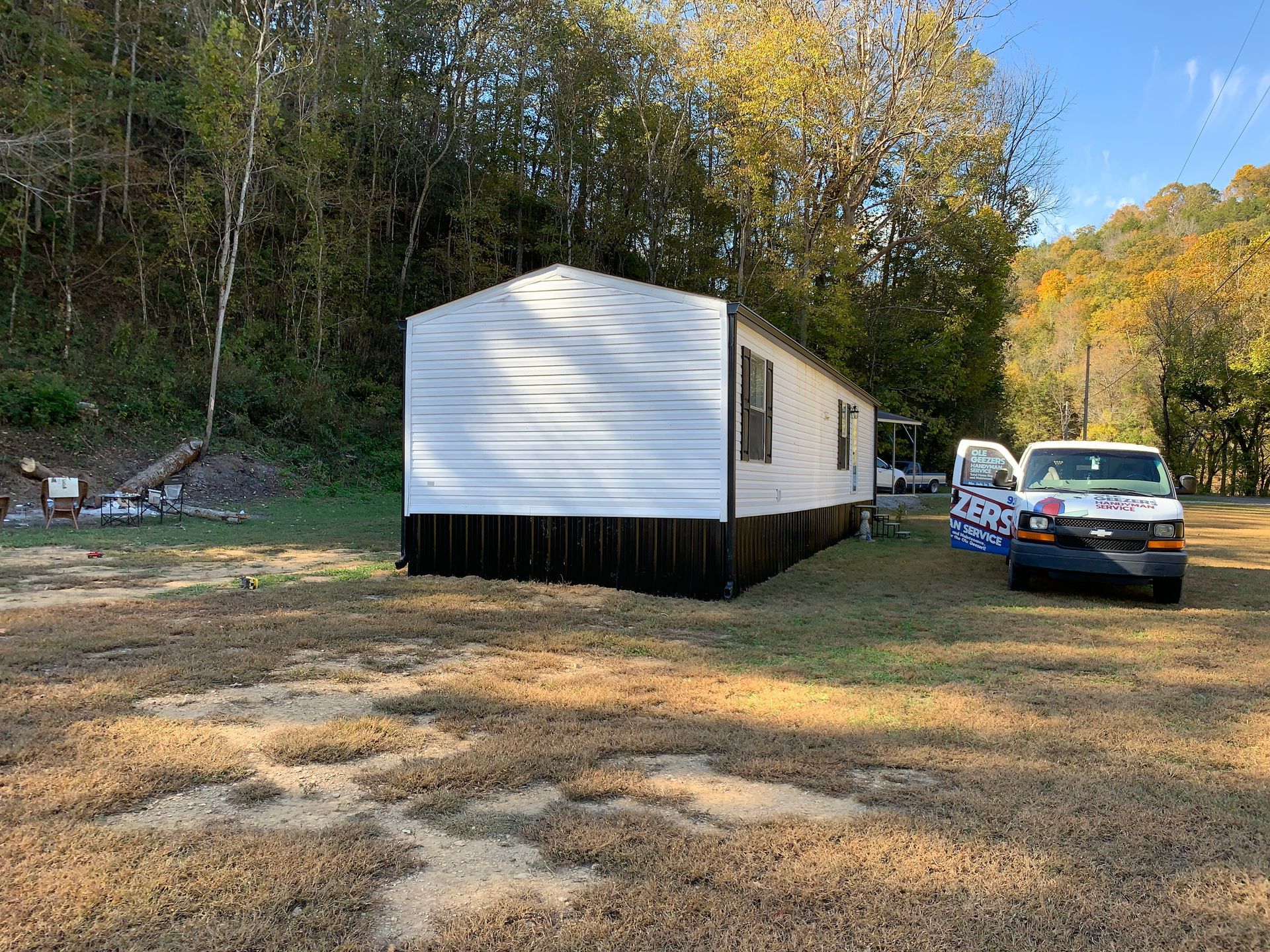 Mobile home with white siding and black trim, van parked next to it, set in a yard with trees in the background.