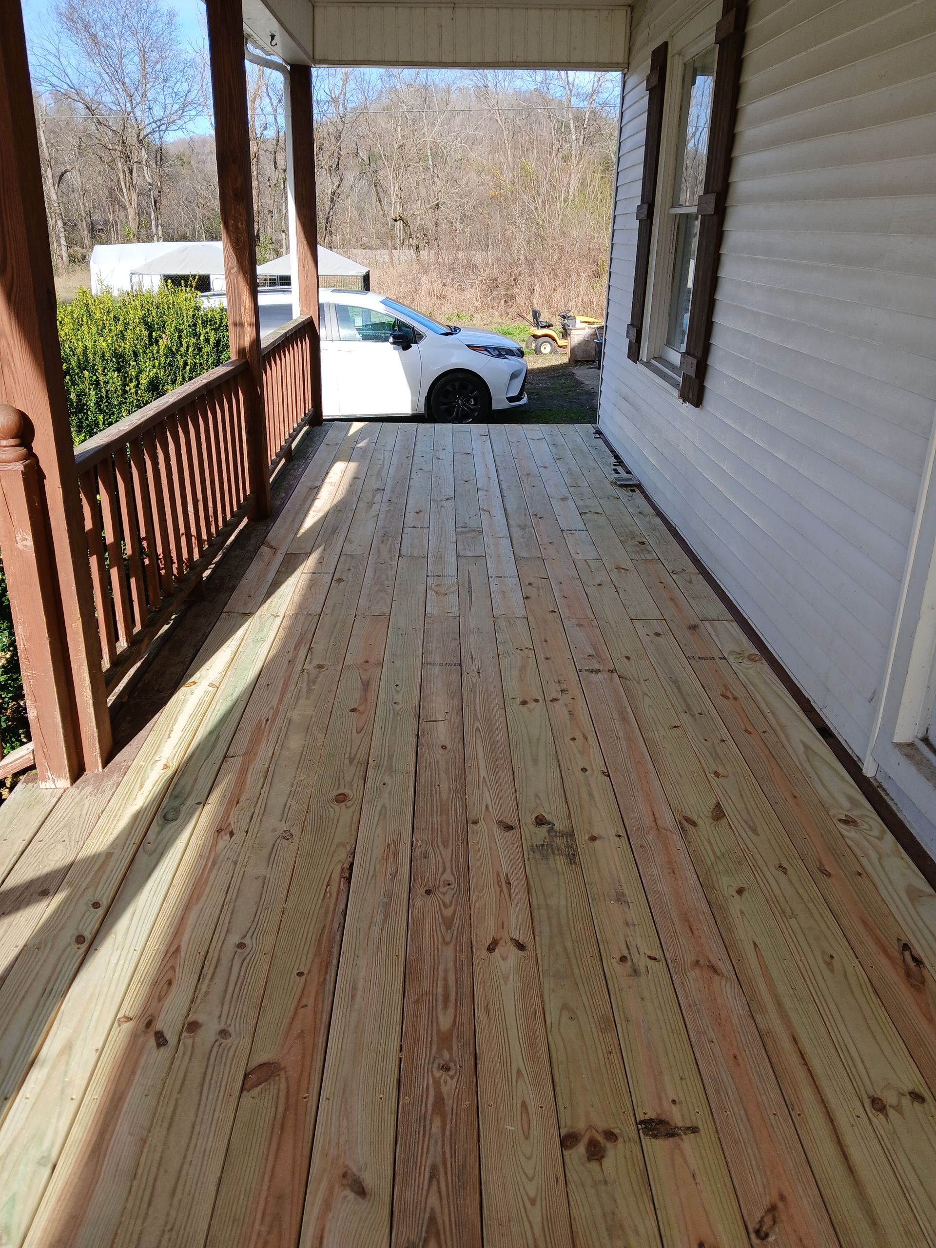 Wooden porch with a brown railing, white siding, and a parked white car in the background.