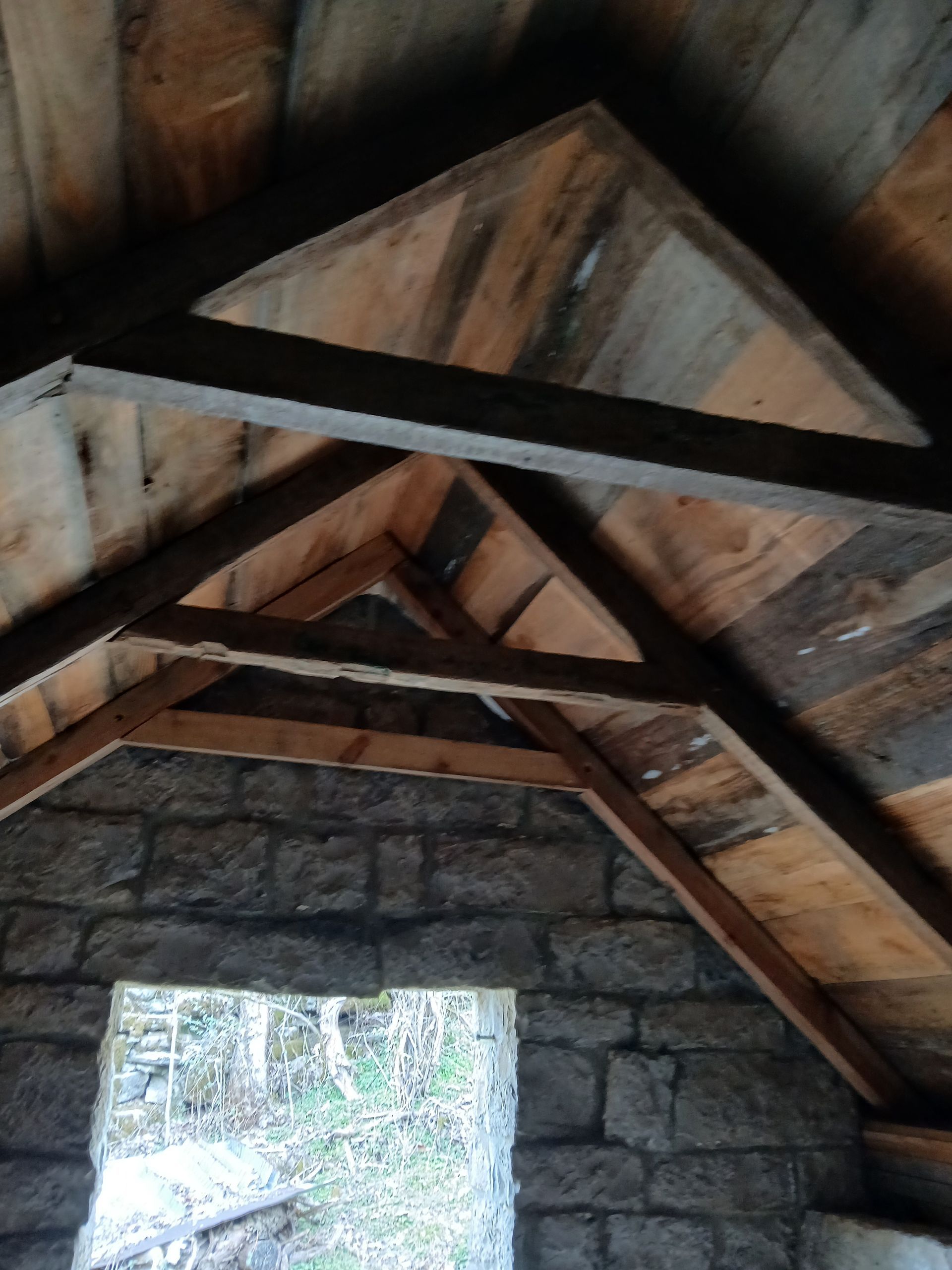 Interior view of a rustic building with wooden beams supporting a peaked ceiling. Stone walls surround a doorway.
