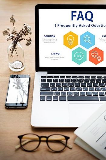 Laptop displaying FAQ page next to a cracked phone, glasses, and flowers on a wooden table.