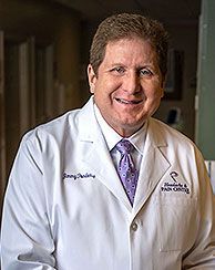 Smiling man in white lab coat, purple tie, at the Headache & Pain Center.