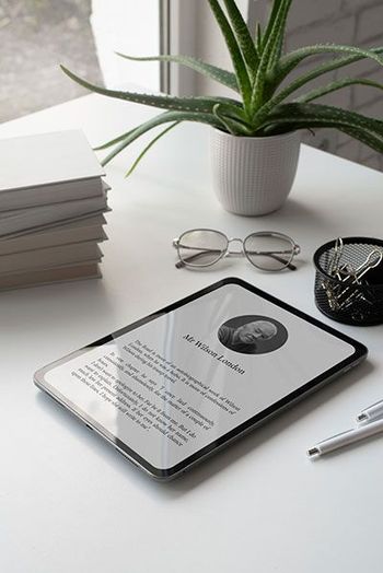 Tablet on a white desk displays a document with a photo and text, next to a stack of books, glasses, aloe plant, and pen.