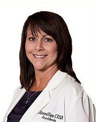Woman with brown hair wearing a white lab coat, smiling. Text on coat reads,