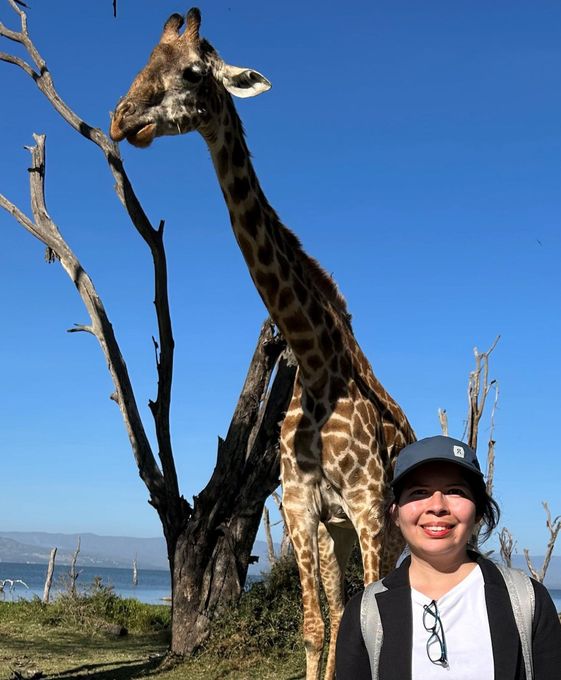 Woman smiling in front of a towering giraffe, with a lake and blue sky in the background.