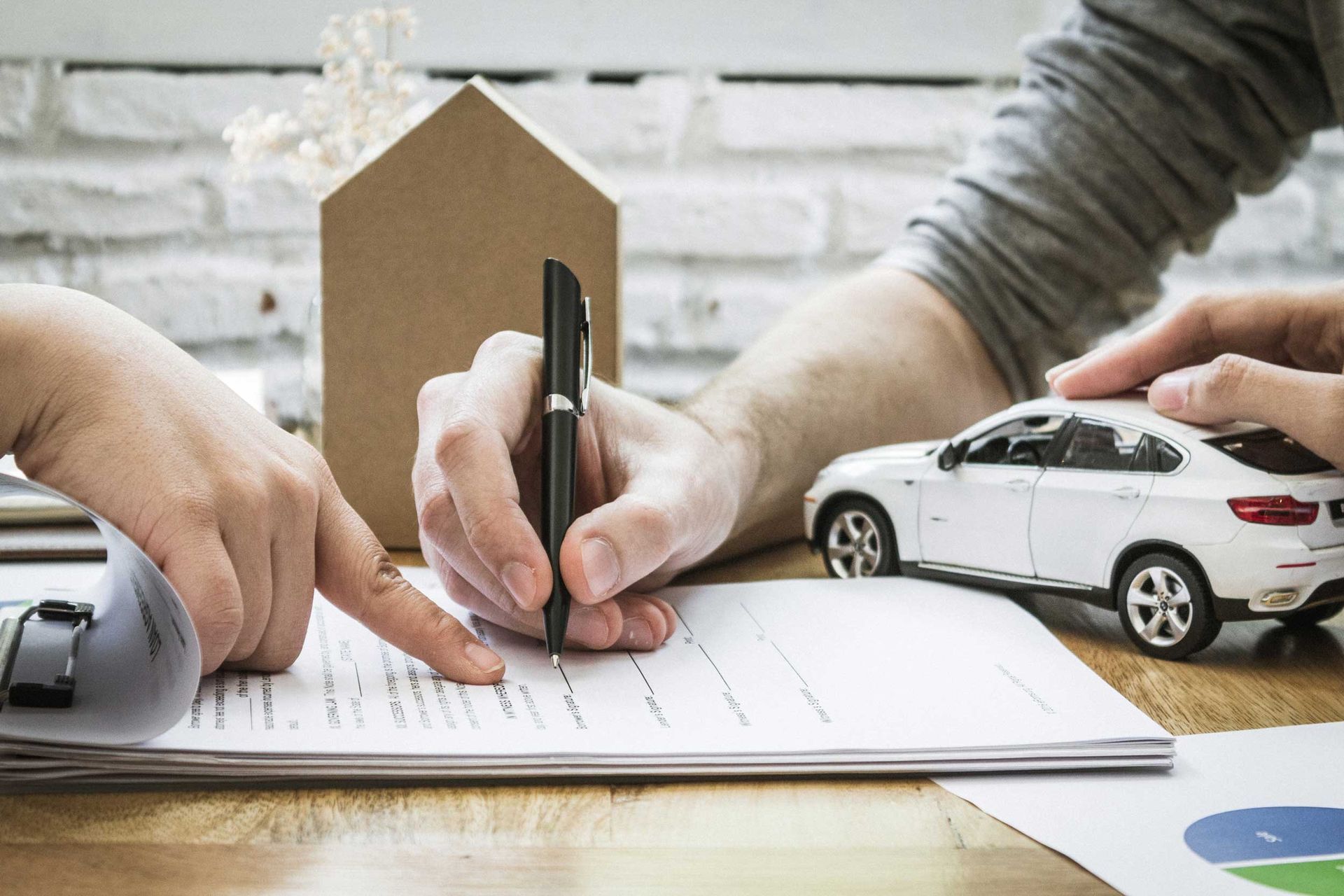 Hands signing a document, with a car model and house model on a table.