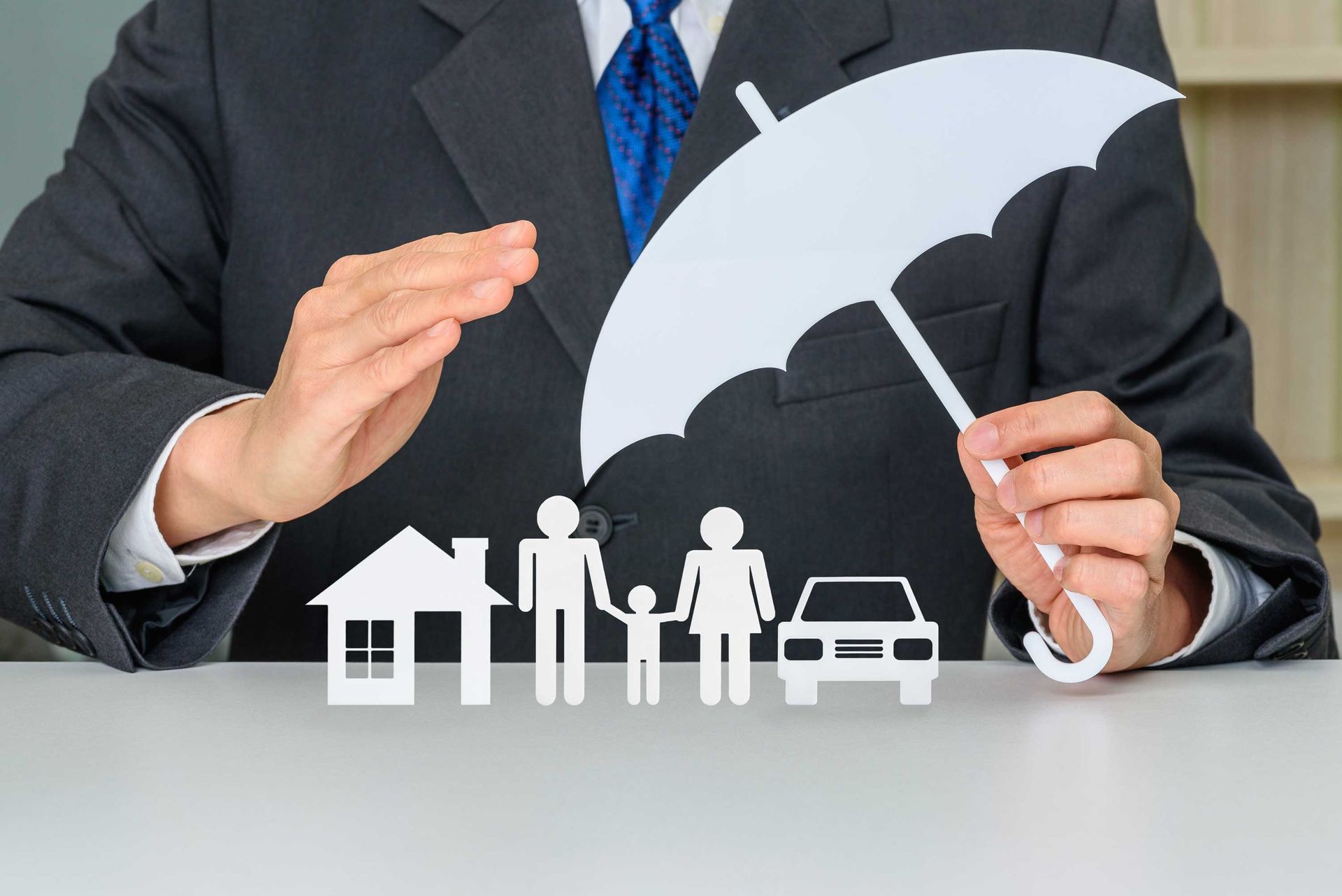 Person in suit holding paper umbrella over family, house, and car cutouts, symbolizing insurance. Person in suit holding paper umbrella over family, house, and car cutouts, symbolizing insurance.