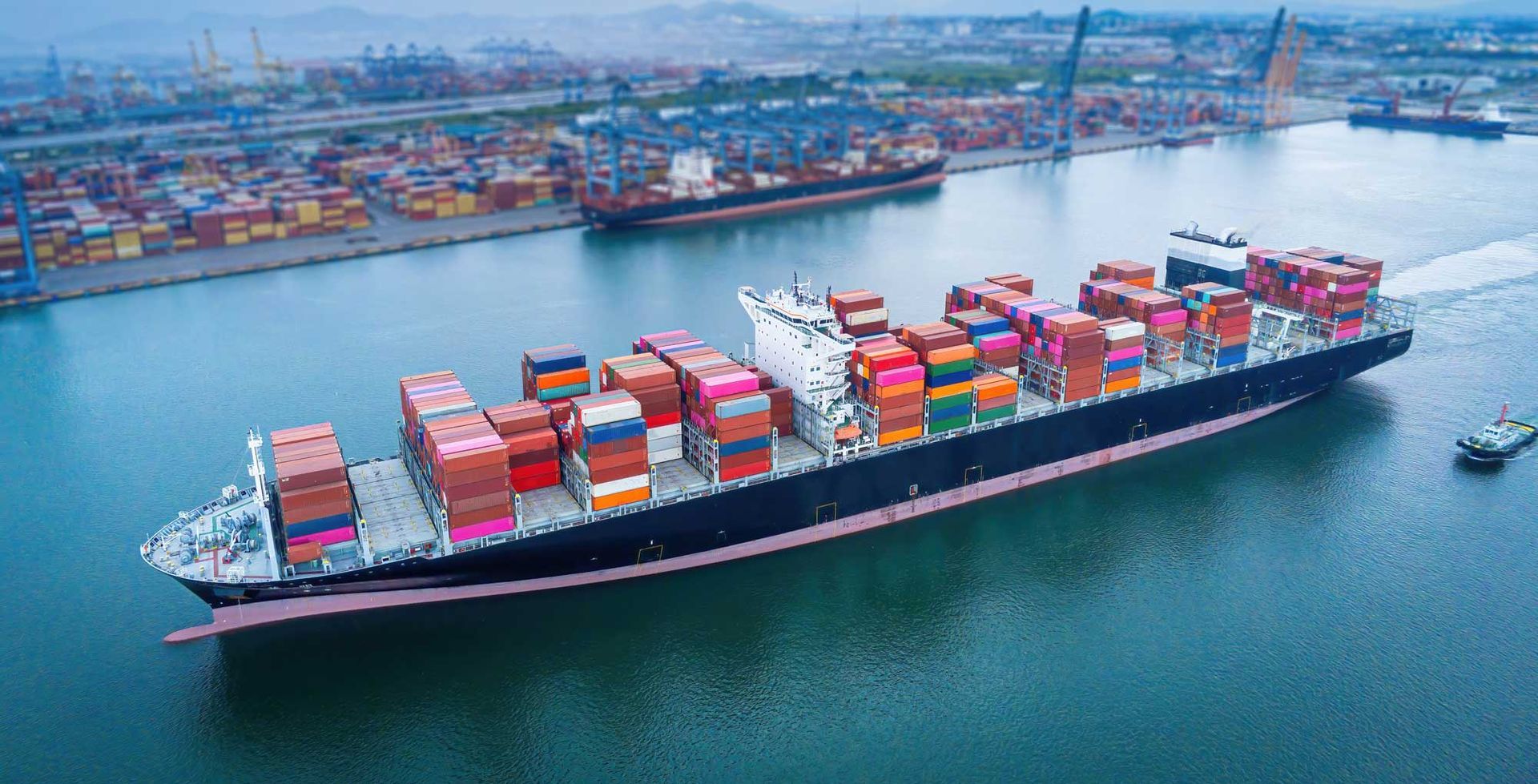 Cargo ship loaded with containers sailing in a harbor, with other ships and cranes in the background.