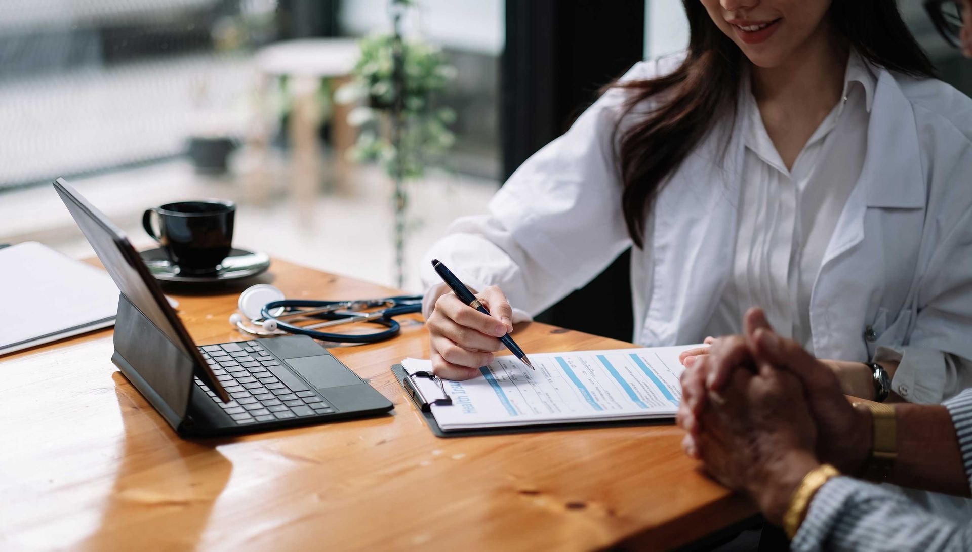 Doctor writing on a form during a consultation with a patient in an office. A laptop and coffee cup are visible.