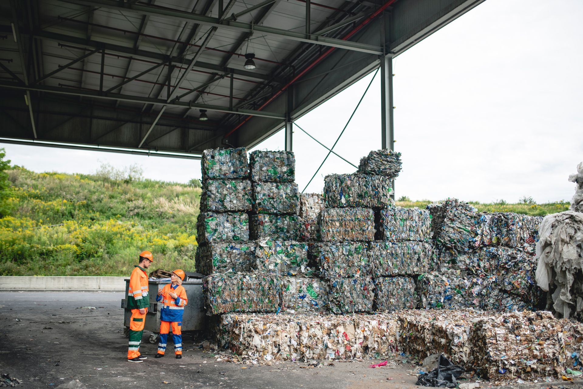 Two men are standing in front of a pile of recycling bales.