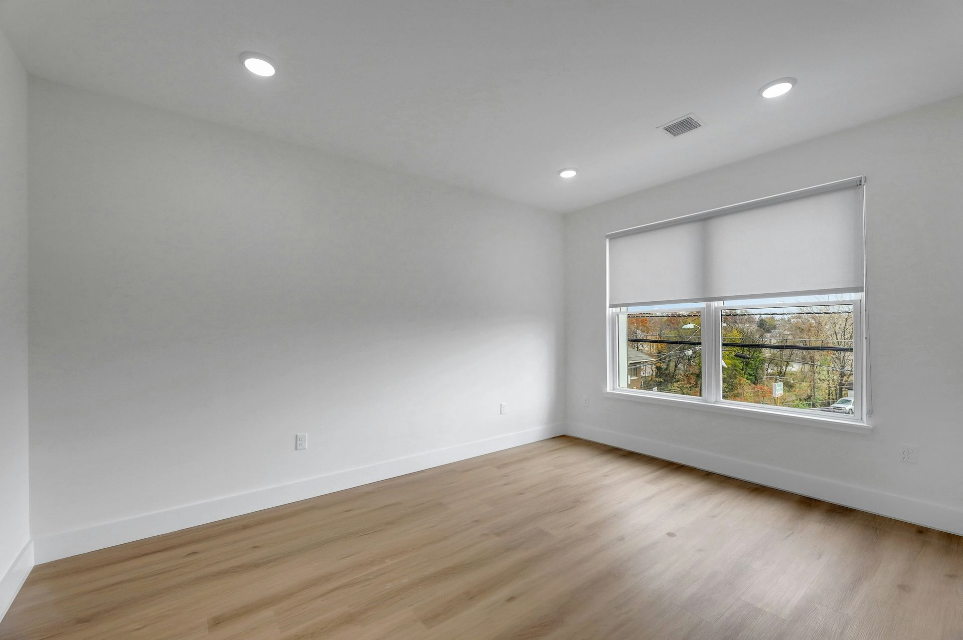Empty room with light wood floor, white walls, window with shade, and recessed lighting.