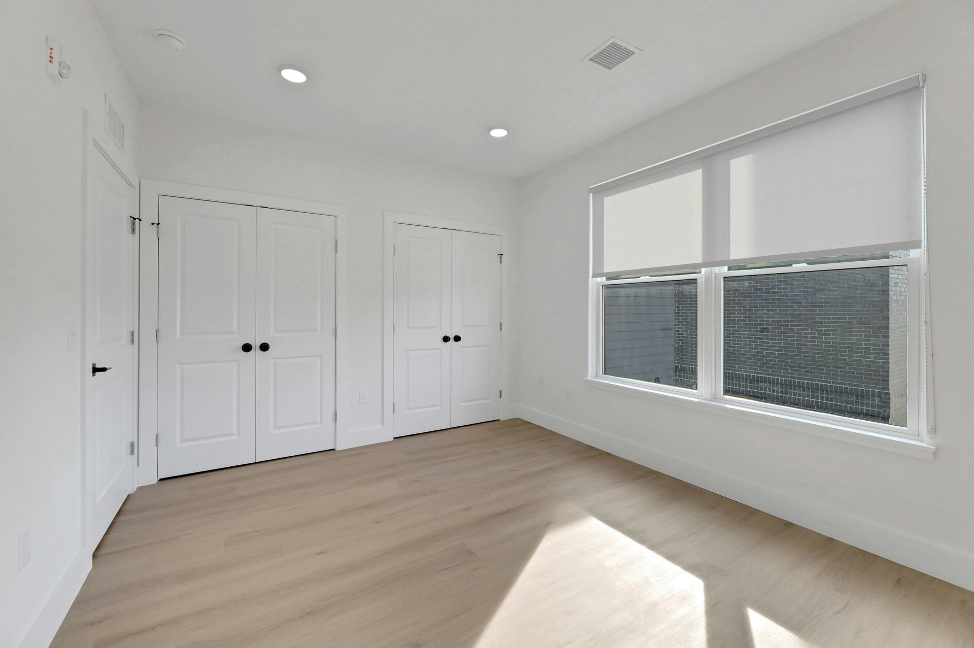 Empty, white-walled room with two doors and a window. Light wood floor, white blinds, and two recessed lights.