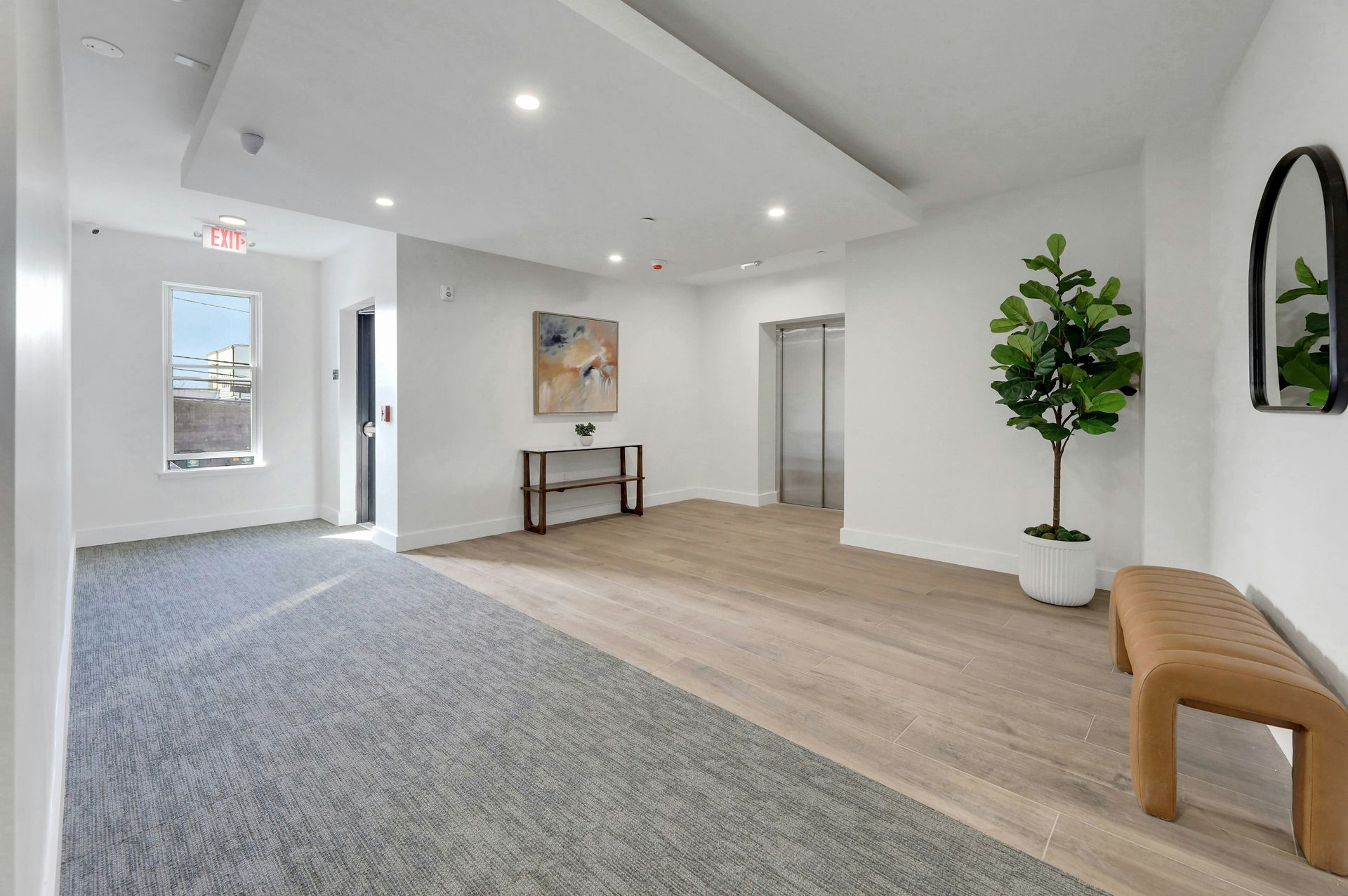Hallway with carpet and hardwood floors, white walls, art, a bench, a potted tree, and an elevator.
